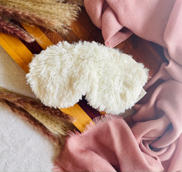 Warmie Plush Eye Mask styled on a wooden block surrounded by a blush scarf and fall twigs