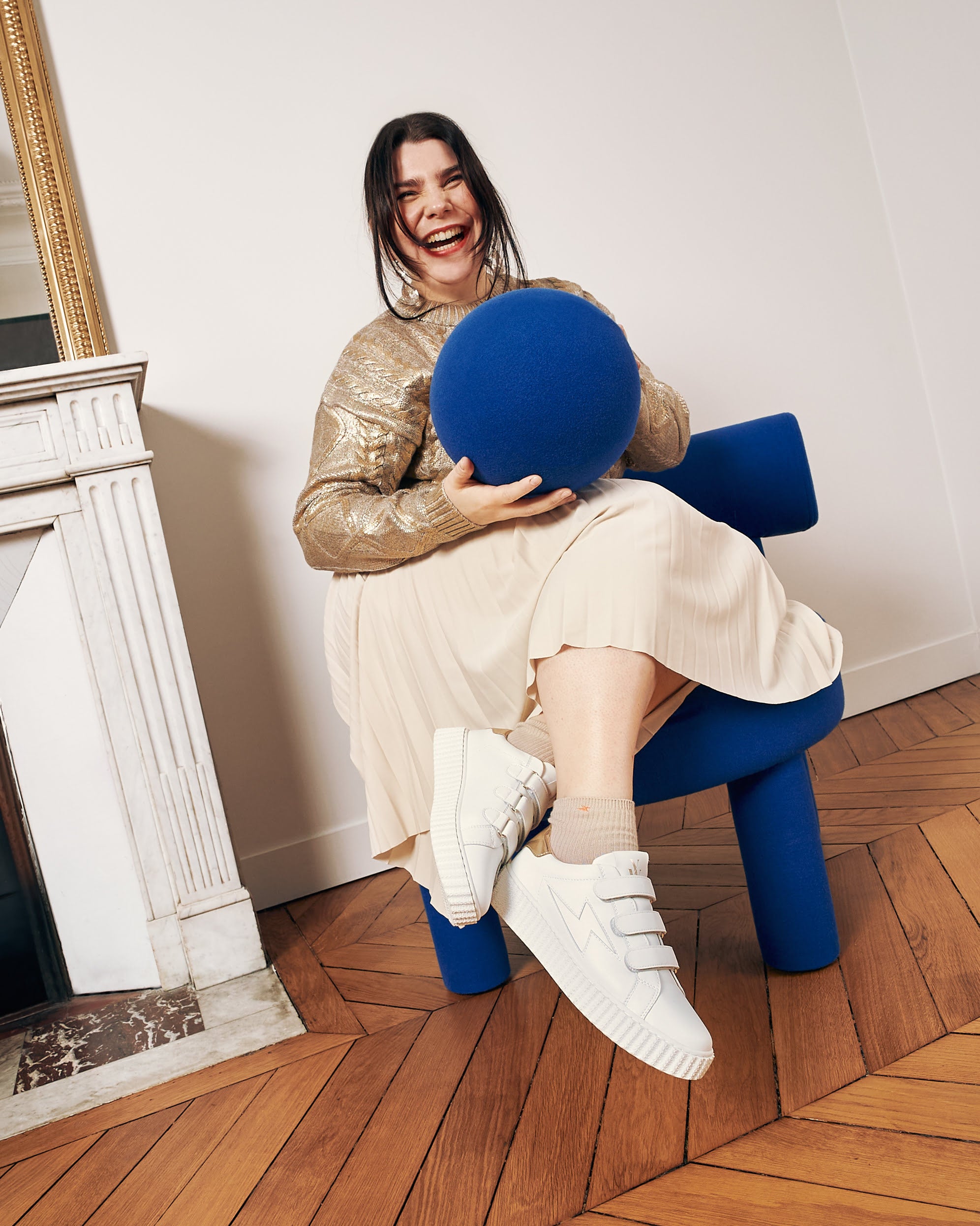 Young woman wearing Maria white and gold storm sneakers with velcro, sitting on a blue chair.