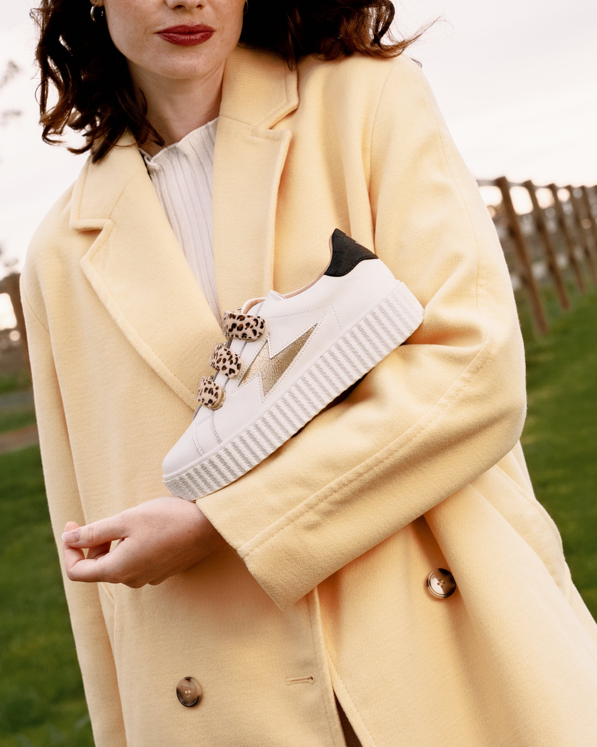 Young woman wearing a yellow coat with Vanessa Wu white and gold Maggie storm sneakers with leopard velcro straps.