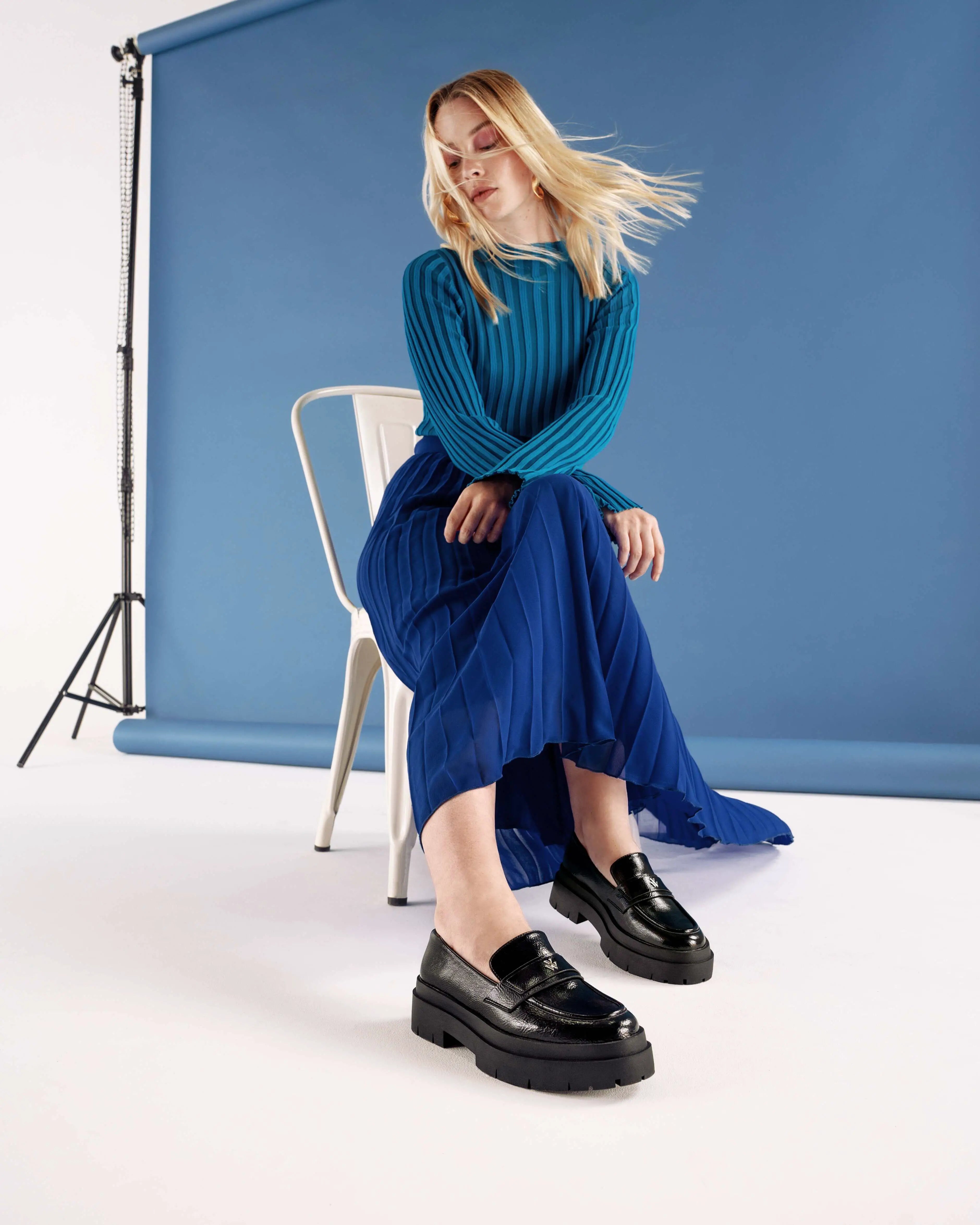 Young woman wearing a blue outfit and the black Rosalie loafers with a notched platform, sitting on a chair.