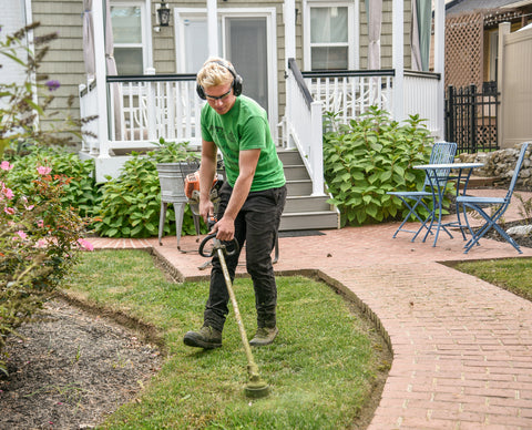 A young man with headphones on is using a weed wacker outside on a front lawn. The weed wacker has zip-ties in it, a popular weeding hack.