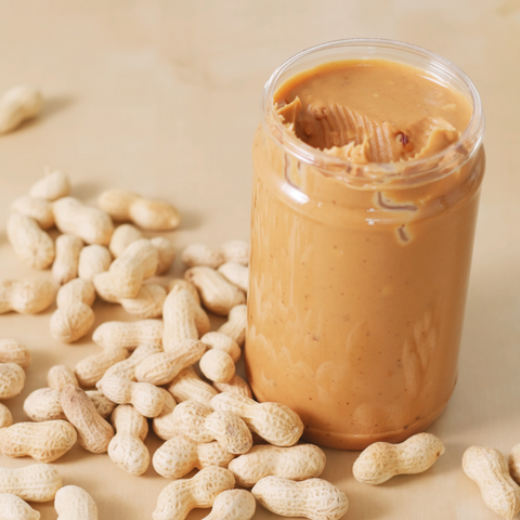 A spoon and jar of smooth peanut butter with shelled peanuts all around it. The peanuts are dispersed all around the jar and come in a variety of light and dark brown tones.