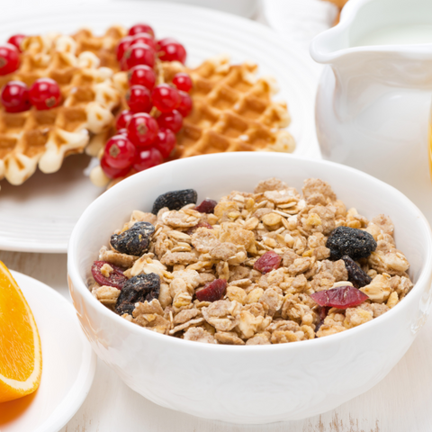 a plate with a warm waffle cooked with muesli grains and garnished with a variety of vibrant fresh fruit, such as pieces of raspberries, blueberries, and kiwis. A drizzle of maple syrup and a dollop of whipped cream are also added to the waffle.