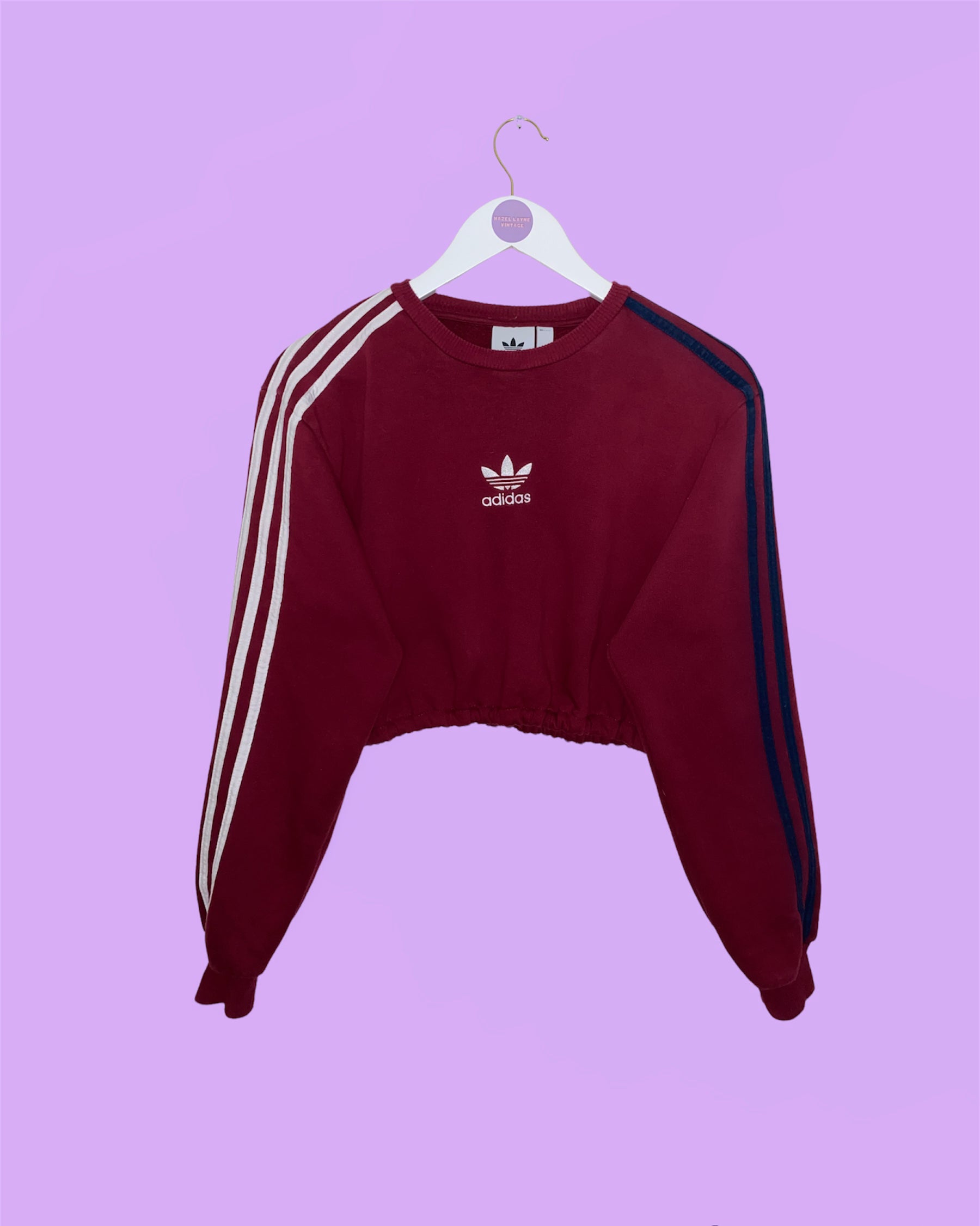 burgundy cropped sweatshirt with white adidas logo shown on a white clothes hanger on a lilac background