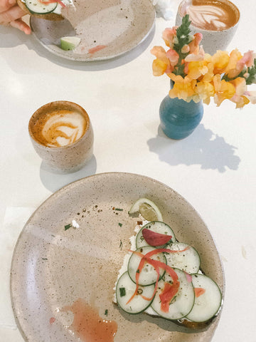 An aerial view of a white table with two plates of cucumber sandwiches, a small vase of flowers, and ceramic mugs of coffee.