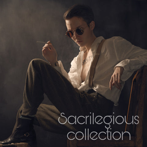 Foto i film noir stil av en androgyn person i vit skjorta och mörka byxor som sitter på ett par resväskor, röker en cigarett och bör ett par runda solglasögon med crucifix på. Fotograf John Stenberg