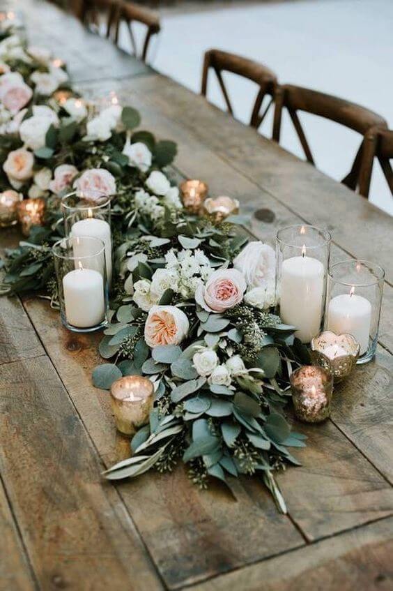 Hidden Botanics elegant flower and greenery table centerpiece with white candles in glass holders, set on a rustic wooden table for wedding decor.