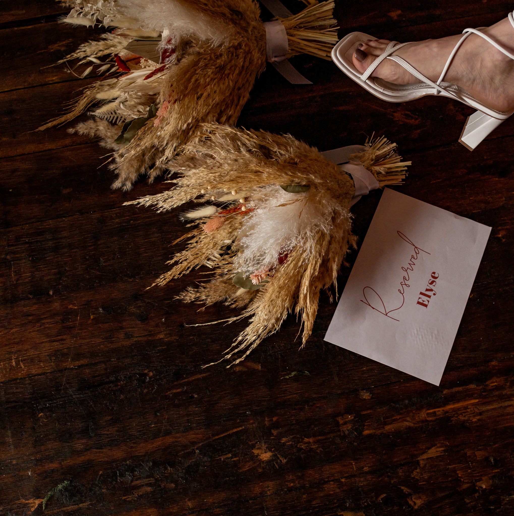 A wedding bouquet of dried flora by Hidden Botanics rests on a wooden floor beside a ’Reserved’ sign with elegant calligraphy.