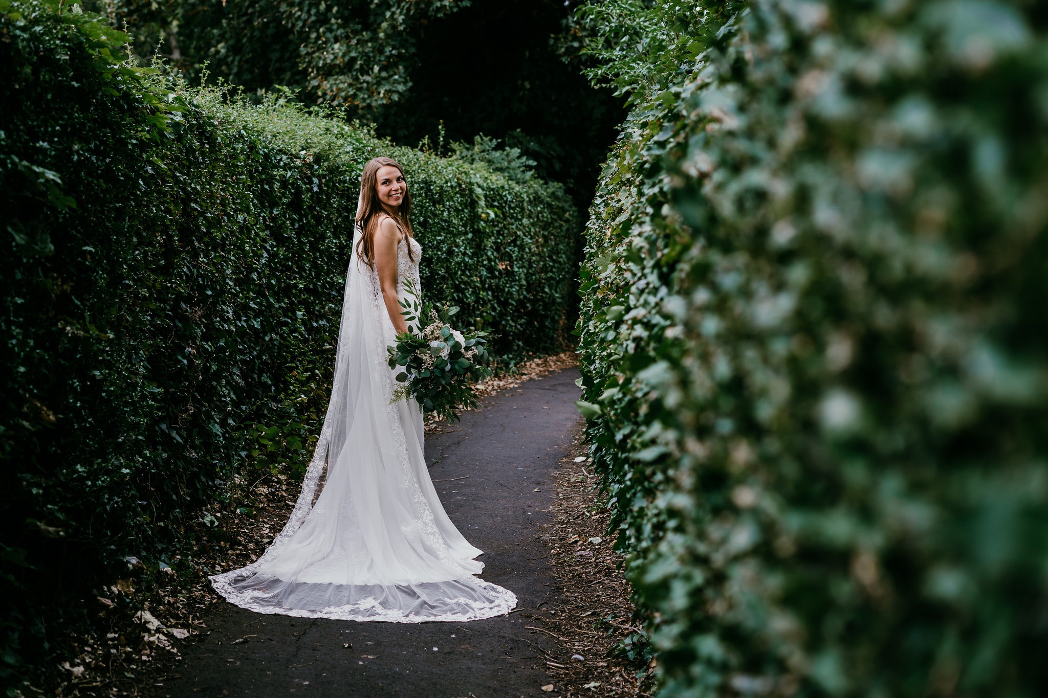 Bride posing with a beautiful bouquet by Hidden Botanics on a hedge-lined path.