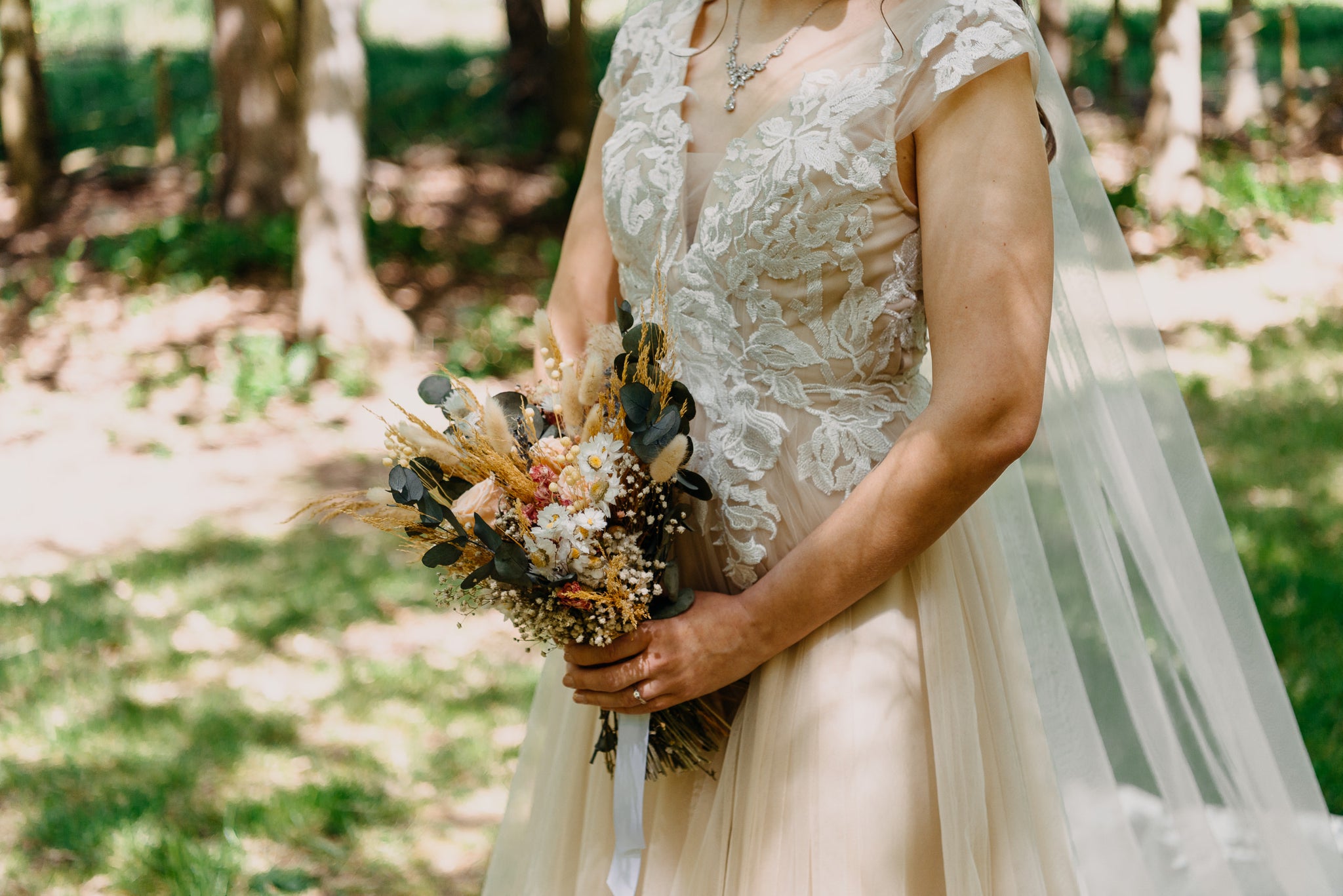 A bride in a lace dress holds a textured Hidden Botanics bouquet featuring dark foliage and white blooms in a serene woodland setting.