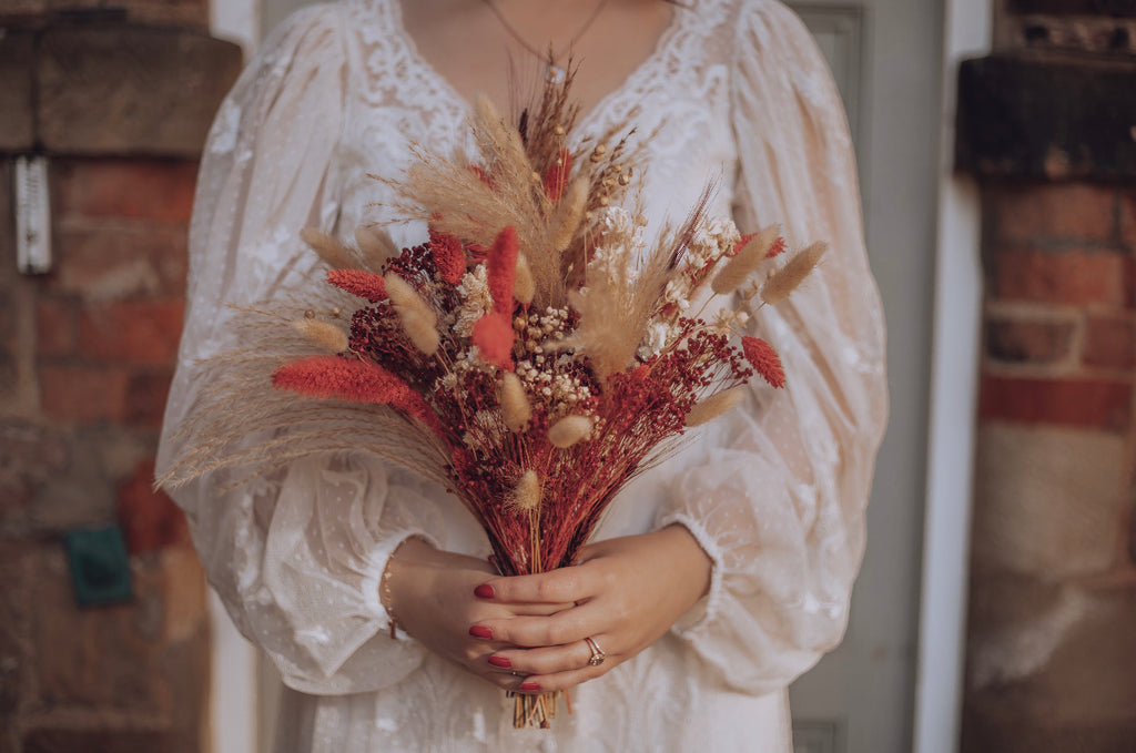 A bride’s hands can be seen holding a burnt orange dried flower bouquet by Hidden Botanics.