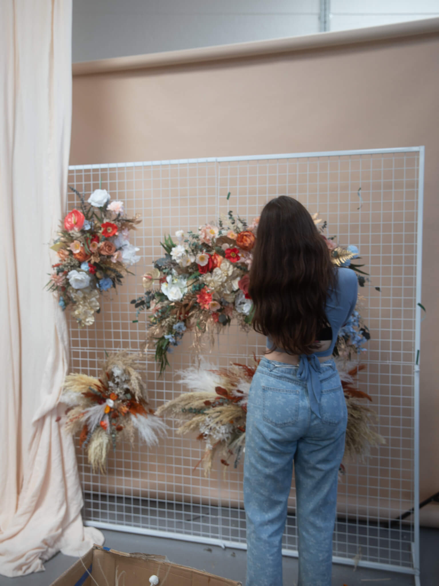 A person views an array of vibrant dried floral arrangements on a grid wall, showcasing an assortment of flowers and foliage in different colors and textures.