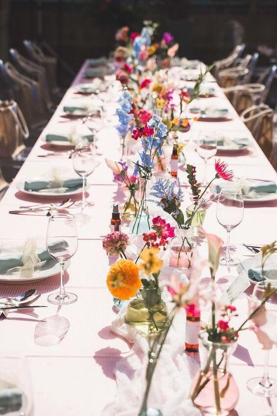 Vibrant and colorful flowers arranged in small glass vases down the center of a long outdoor dining table, set for a festive occasion in bright sunlight.