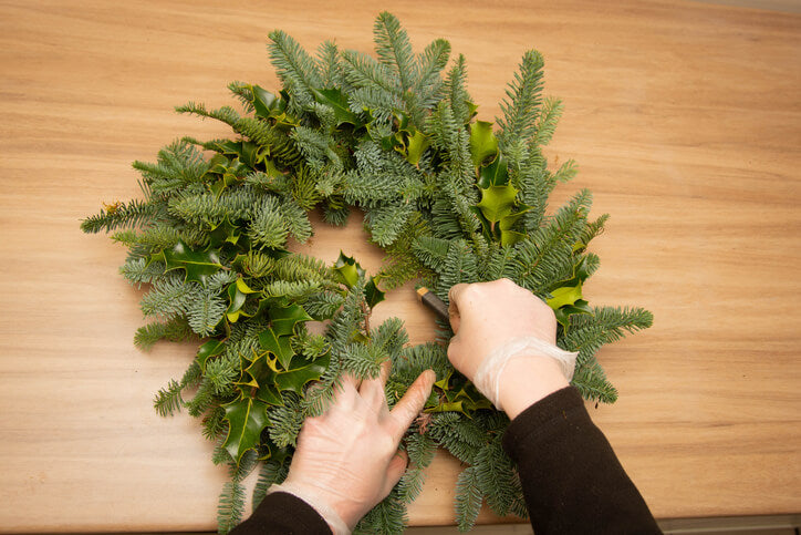 A person's hands are shown crafting a wreath with various evergreen branches, including fern and holly, on a wooden surface.