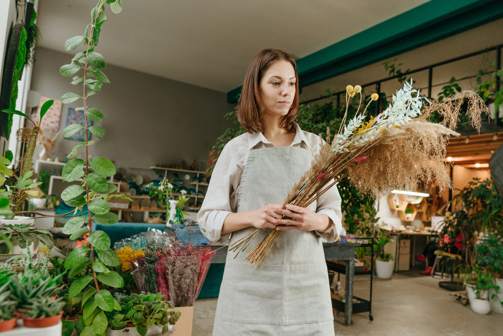 A florist is surrounded by plants as she holds dried flower stems.