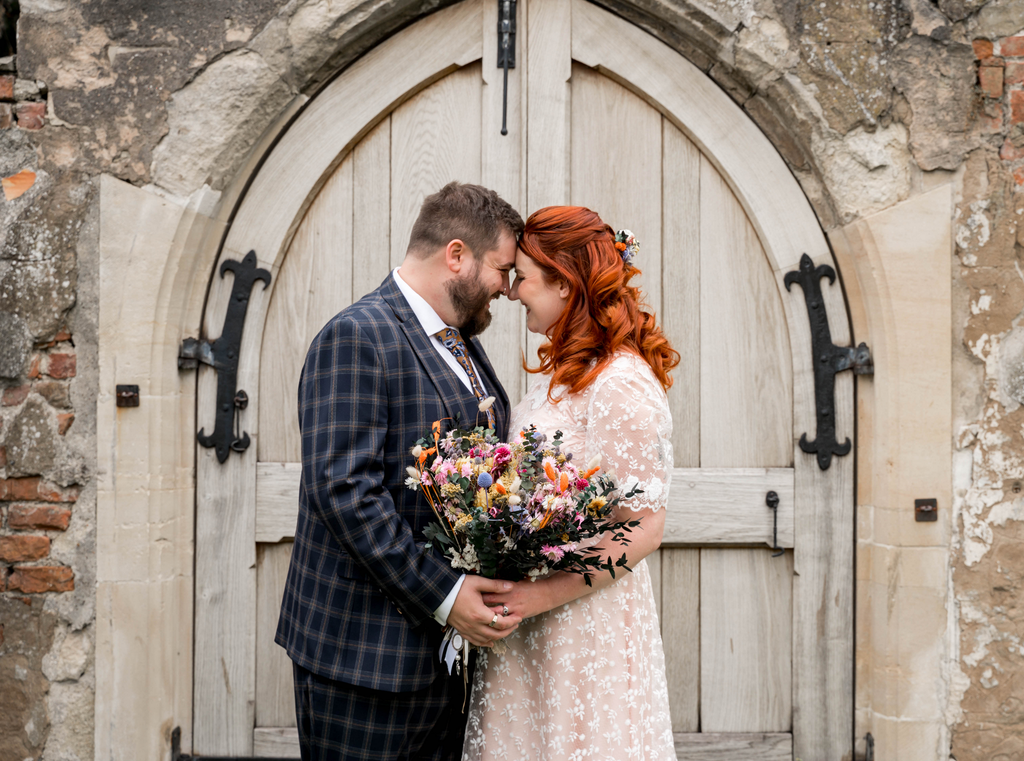 A bride and groom stand together in front of a wooden doorway, holding a wedding bouquet from Hidden Botanics.