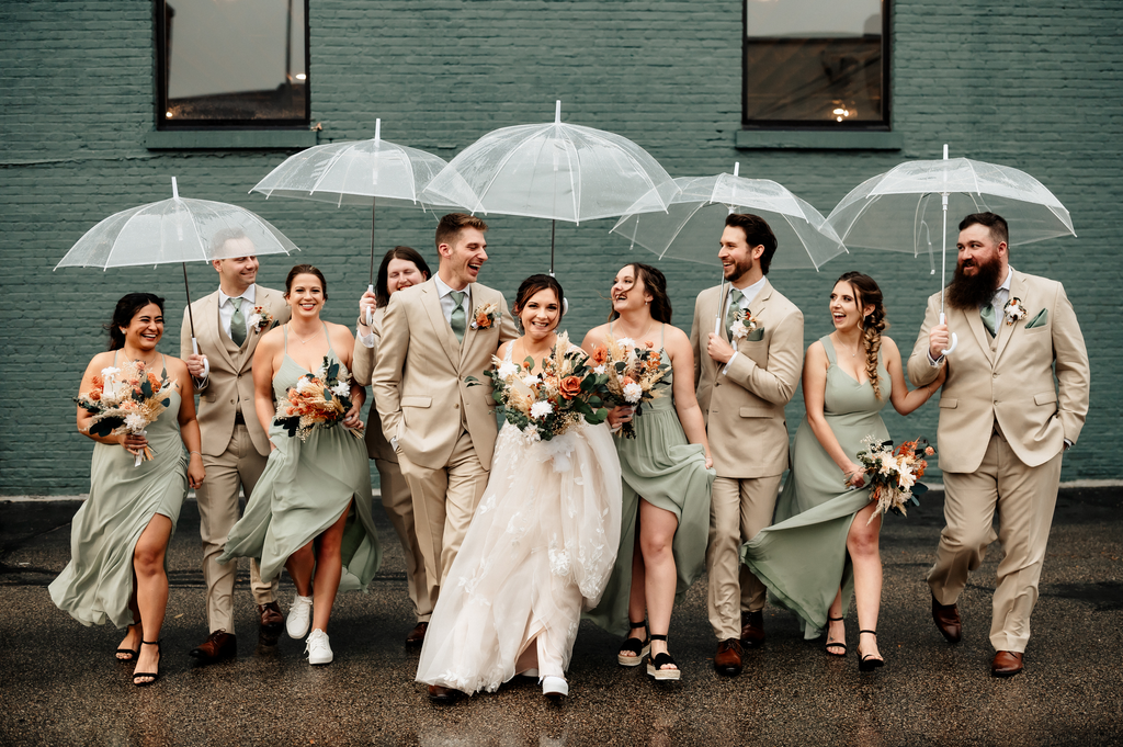 A bridal party wears beige and green outfits, holding clear umbrellas and dried flower bouquets.