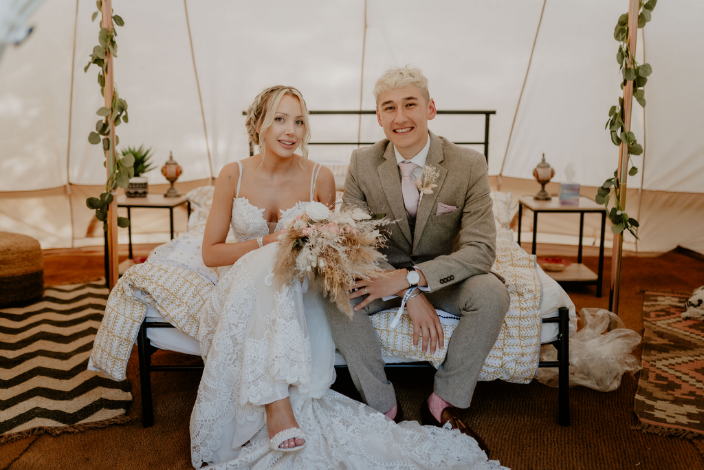 A bride and groom sit inside a tent, smiling at the camera, holding a Hidden Botanics’ dried flower arrangement.