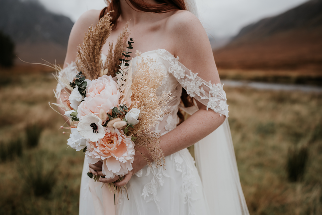 A bride stands in a rural location holding a dried flower bouquet by Hidden Botanics.