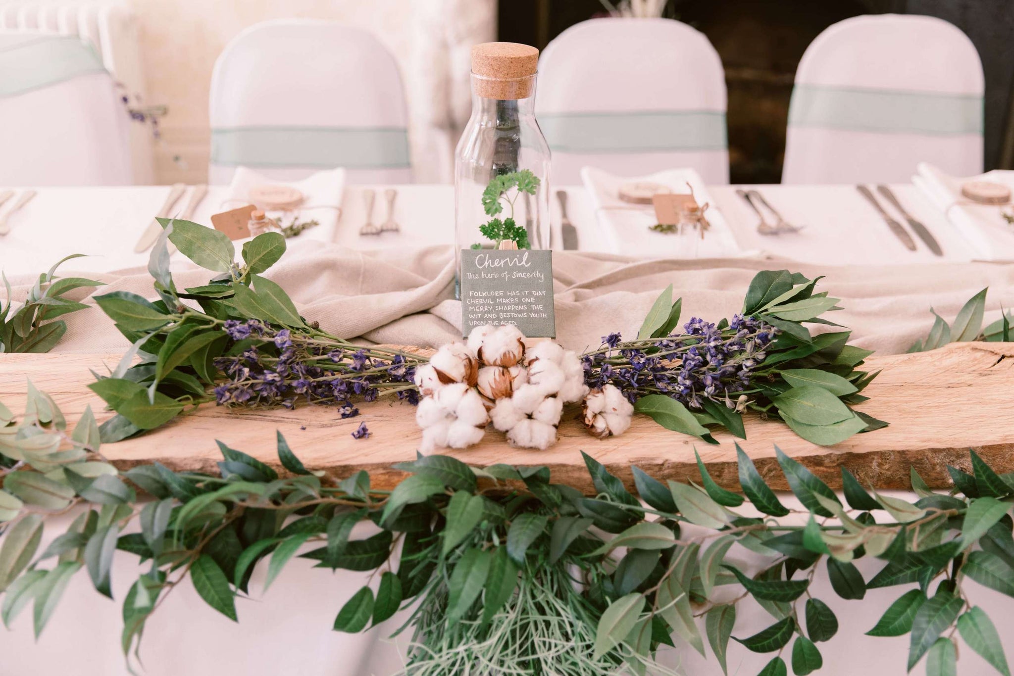 Rustic table decor with a central wooden beam, green eucalyptus leaves, cotton stems, and lavender, accompanied by a personalized bottle centerpiece.
