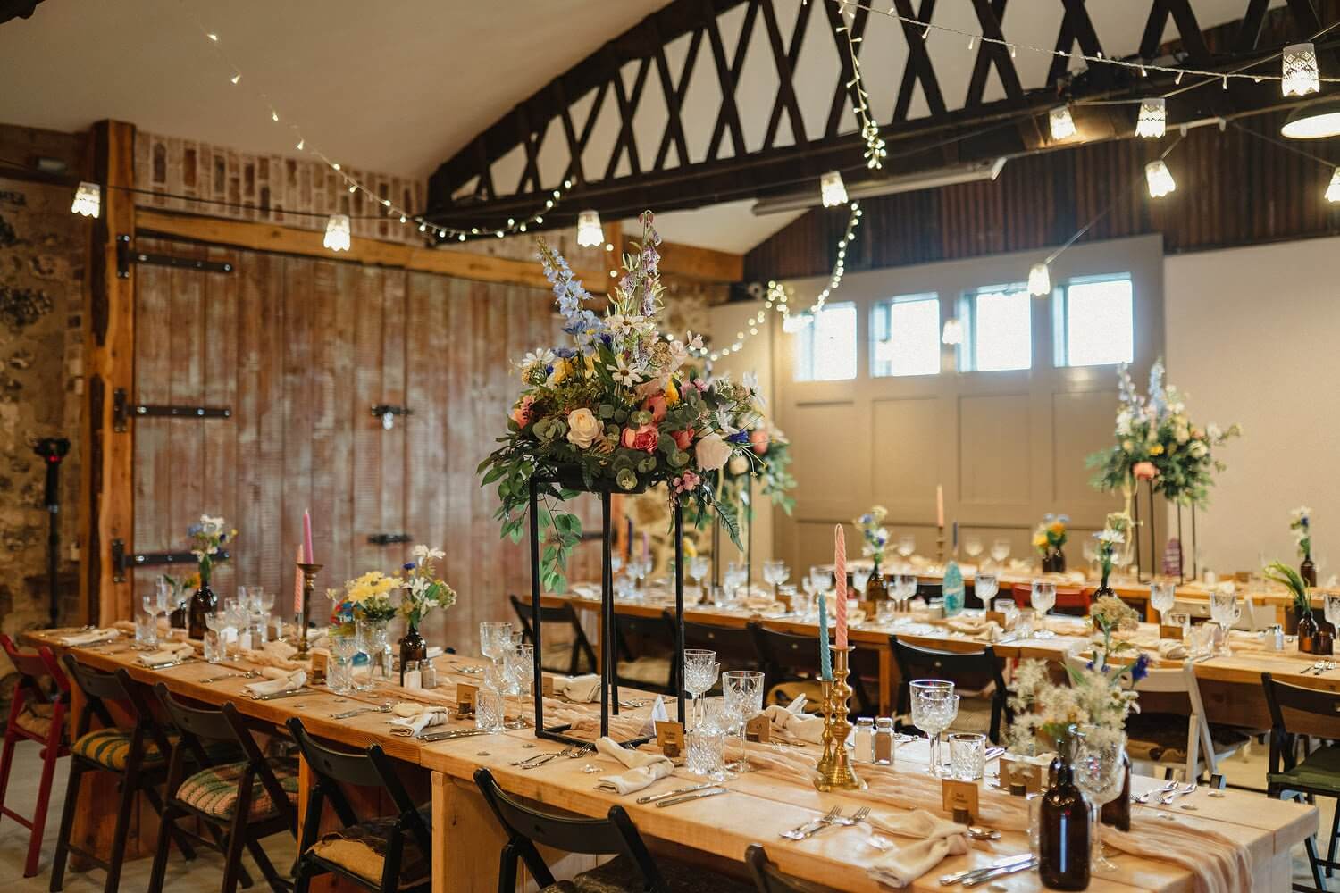 Rustic barn wedding reception with elegantly set long wooden tables, tall floral centerpieces, and string lights, creating a cozy and inviting atmosphere.