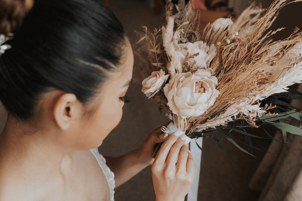 A bride adjusting her wedding bouquet with a mix of artificial and dried flowers.