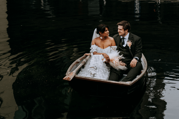 A bride and groom in a boat on the lake holding a cascading bouquet of dried peach and white flowers.