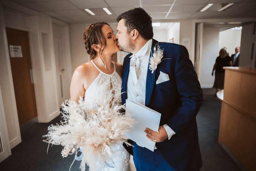A newlywed couple sharing a kiss in a corridor with a white artificial bridal bouquet and matching Boutonniere.