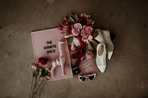 Artificial mixed bridal bouquet with pink orchids, white ankle boots, heart sunglasses, and a sign with a pink toy giraffe arranged on the floor.