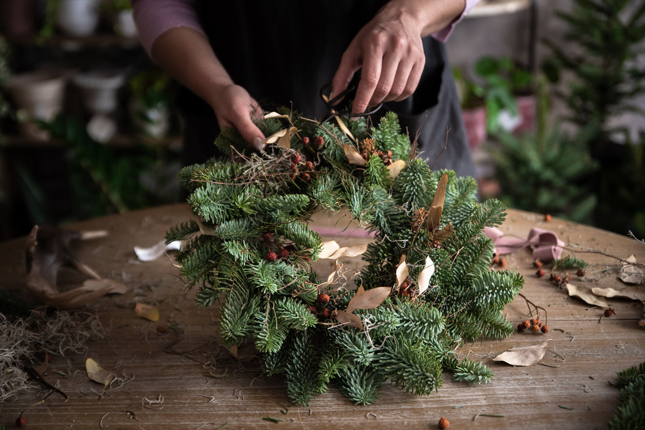 A person's hands are shown trimming and arranging a festive wreath made of evergreen branches, adorned with red berries and dried leaves.