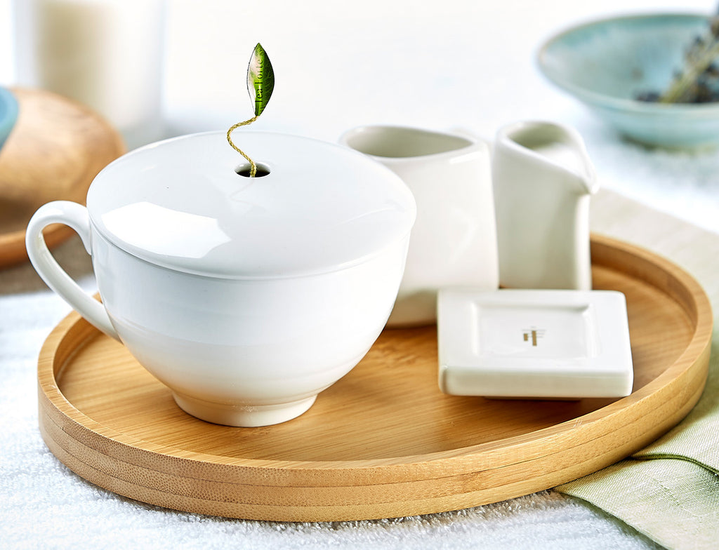 Close up of a white café cup filled with tea next to a tea infuser on a white tea tray presented on a bamboo oval tray.