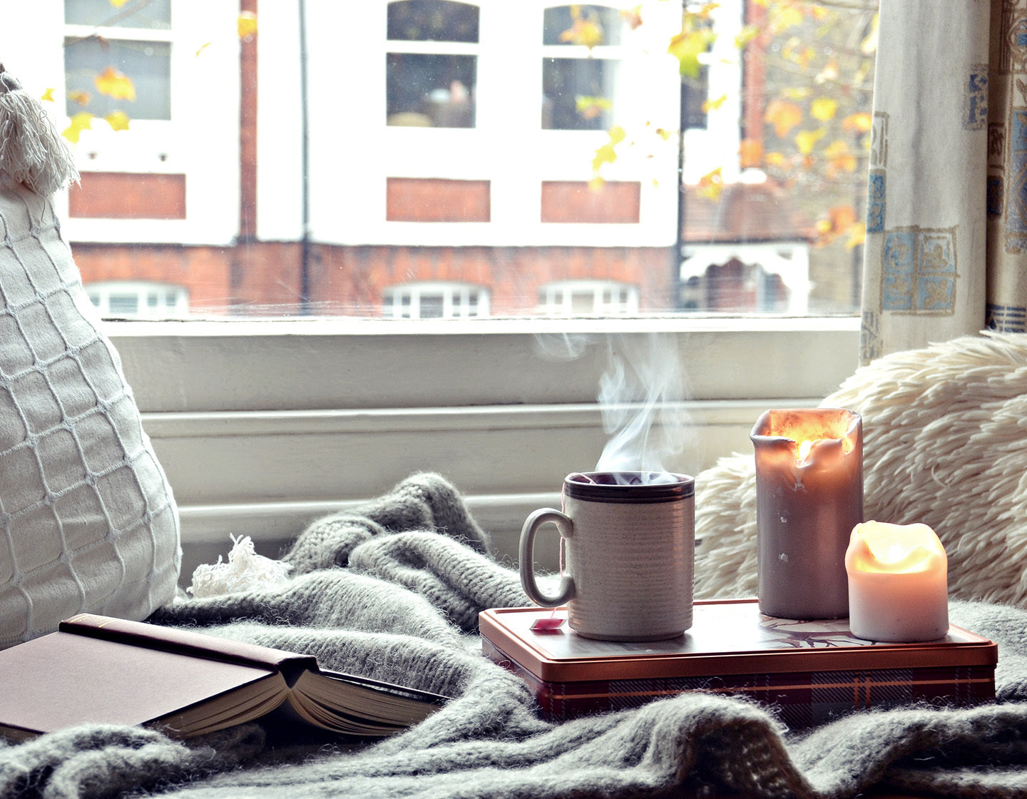 A steaming cup of tea with a book, blanket and lit candle by a window