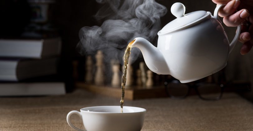 Pouring tea into a white cup from a English white teapot with a chess set and books in the background