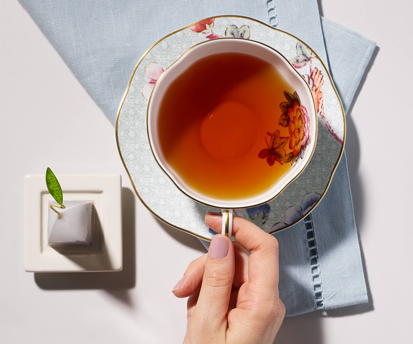 Woman grasping a porcelain cup of tea with a pyramid tea infuser on a tea tray to the side
