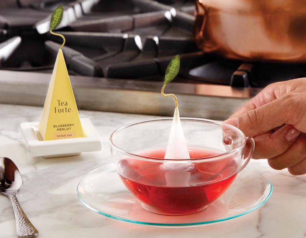 Glass cup and saucer and pyramid tea infuser on a white tea tray.