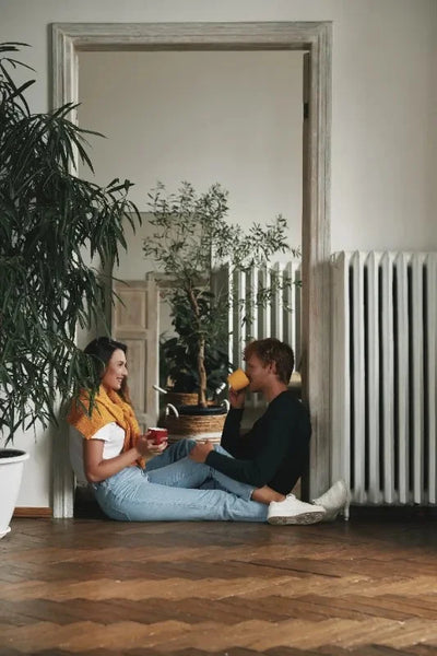 beautiful young couple sitting on a wooden floor in front of a white cast iron radiator and large mirror drinking hot drinks look at each other