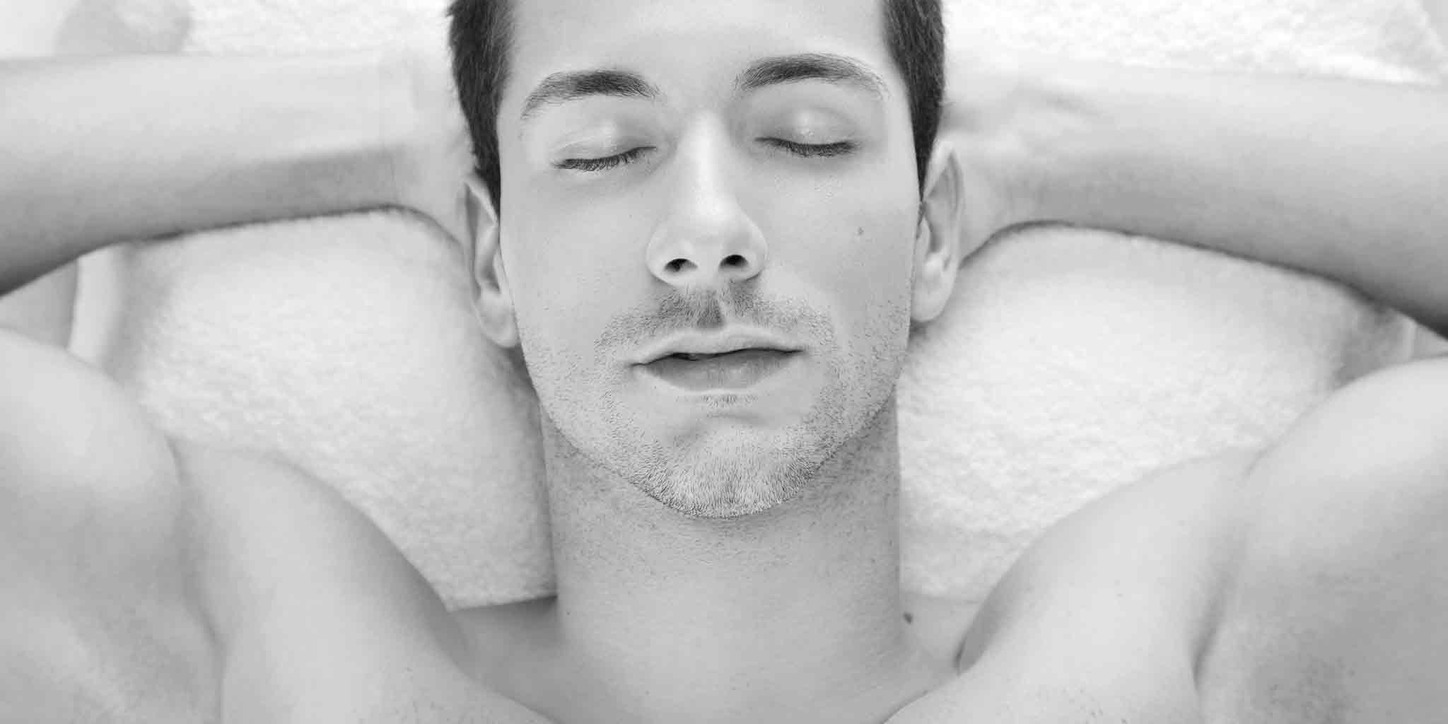 Monochrome image of a relaxed man lying down with eyes closed, embodying the serene experience of a men's massage at Core Benefits Toowoomba, highlighting the anticipated comfort and tranquility.