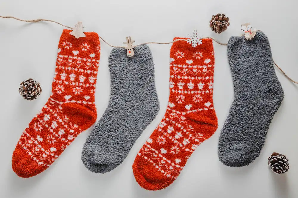 Three pairs of warm fuzzy socks in red and grey colors with Christmas patterns, hanging on a string with wooden clips and decorations, pinecones scattered around.