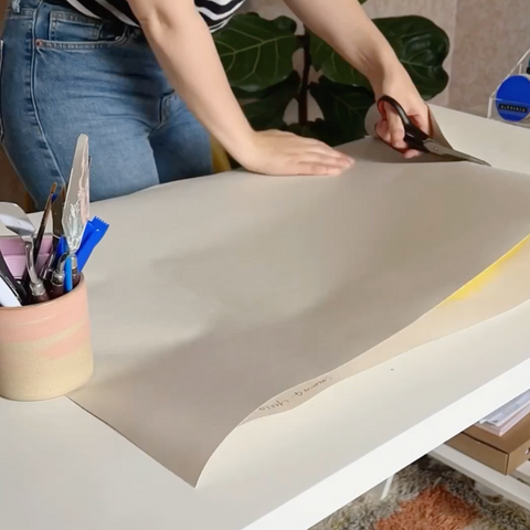 A woman in striped shirt and jeans cutting a big roll of white lining paper placed on a white desk