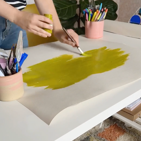 Woman painting with olive green paint onto white lining paper placed on a white desk