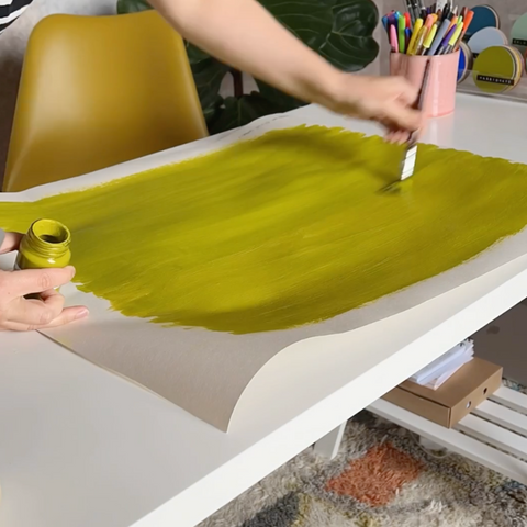 Woman painting with olive green paint onto white lining paper placed on a white desk