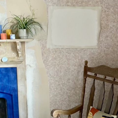 A large sheet of lining paper painted in a neutral white colour is stuck on a wallpapered wall above a wooden chair