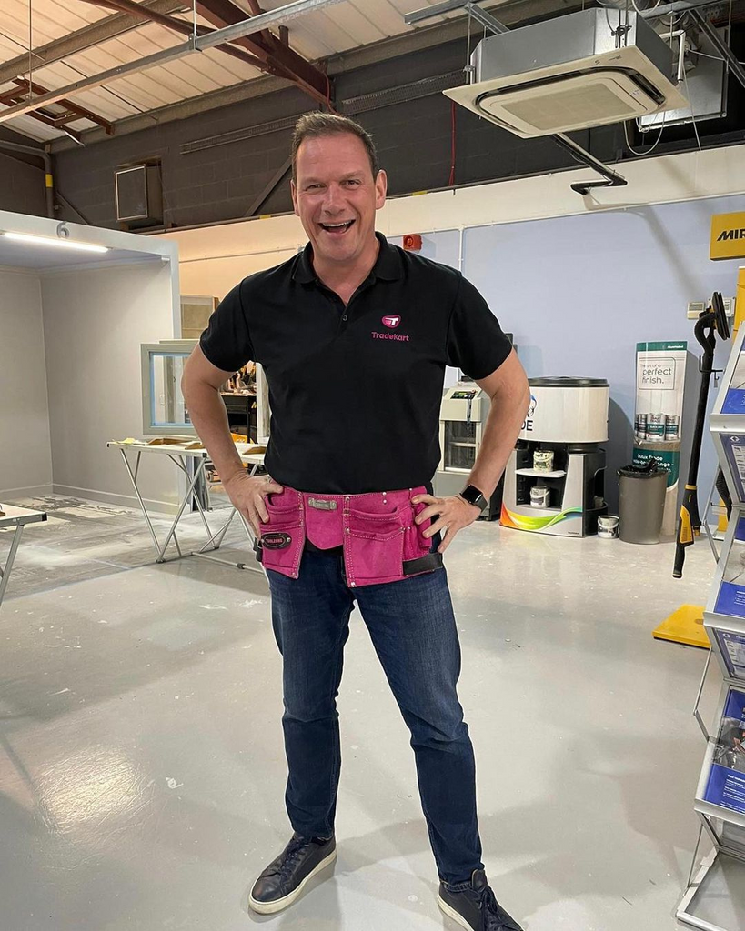 a man with black t-shirt and jeans, photographed in full size, posing with a pink tool belt on