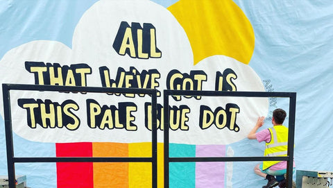 Photo of a man with a bright yellow shirt standing on the bottom right corner of an artwork in a light blue colour, with a rainbow painted in red, orange, yellow, teal and lilac and with a big text with cartoon-like font reading 'all that we've got is this pale blue dot'