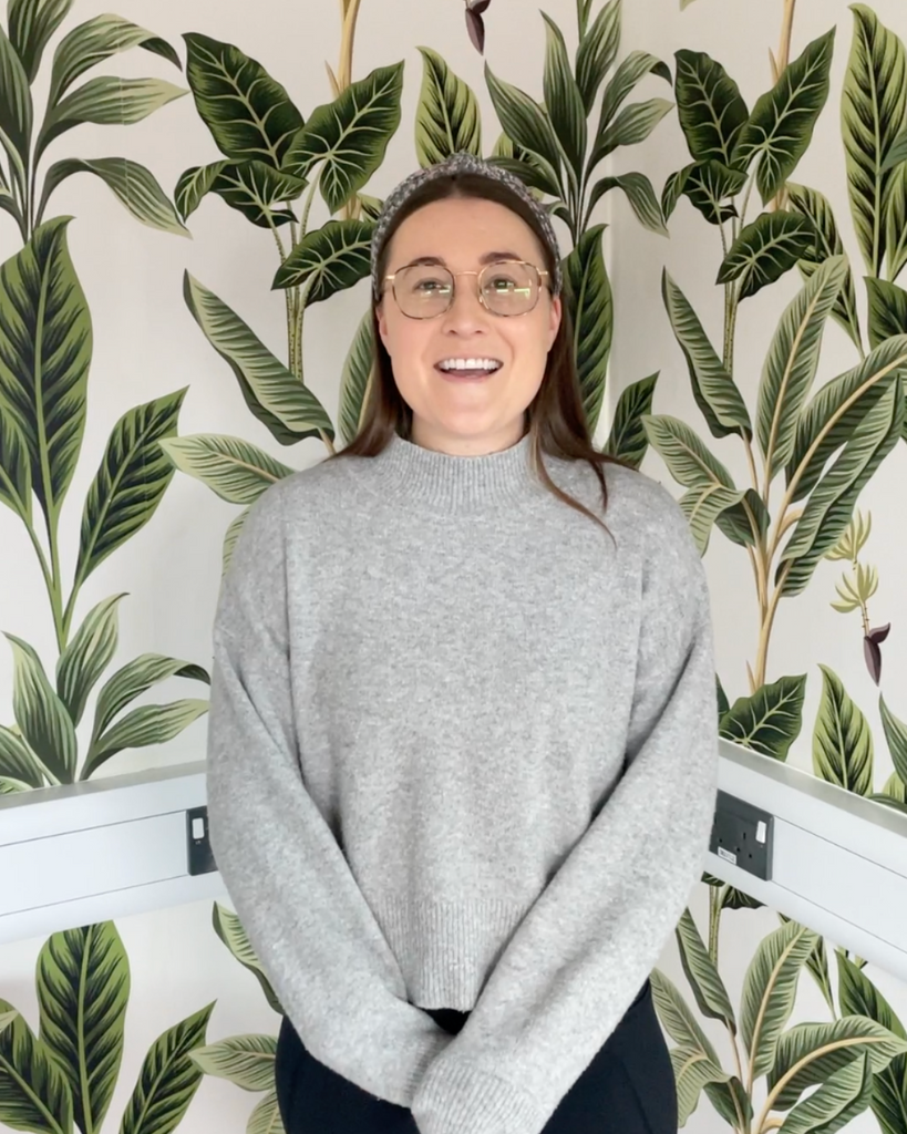 woman with dark blonde hair, glasses and grey sweater, posing in front of a wallpaper in a tropical leaf pattern in white and green