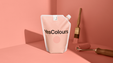 photo of a plastic pouch with logo that reads YesColours positioned in the middle of the frame with a paint brush and paint roller on its right side, all photographed in front of a muted peach colour background