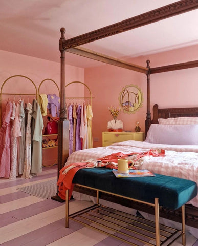 photo of a light pink painted spacious bedroom with a wooden framed bed photographed on the right side, a yellow coloured bedside table on its right and a centred long clothing rack with pastel coloured dresses hanging from it