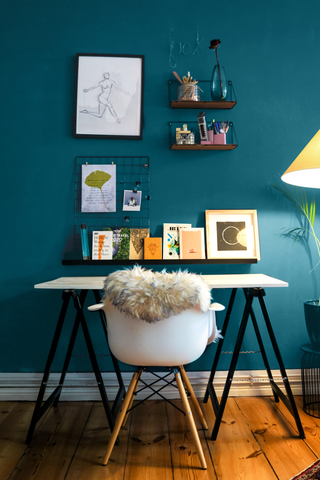 home office interior setting with a wooden desk with metal legs, white chair with a faux sheep skin throw, gallery wall and dark teal painted wall behind the furniture