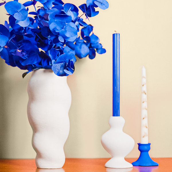 The top of a vintage mid-century wooden sideboard, with placements of a white ceramic vase with blue-painted eucalyptus in it, white ceramic candle holder with blue candle in it, and a small blue-painted candle holder with a tall white candle in it, all photographed in front of a warm creamy white coloured wall 