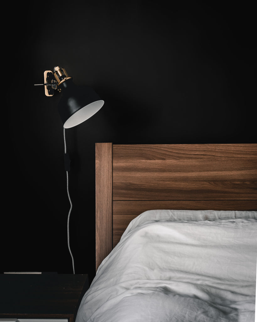 Wooden bed corner with a bedside lamp hanging on a wall next to it, all photographed on a black painted wall